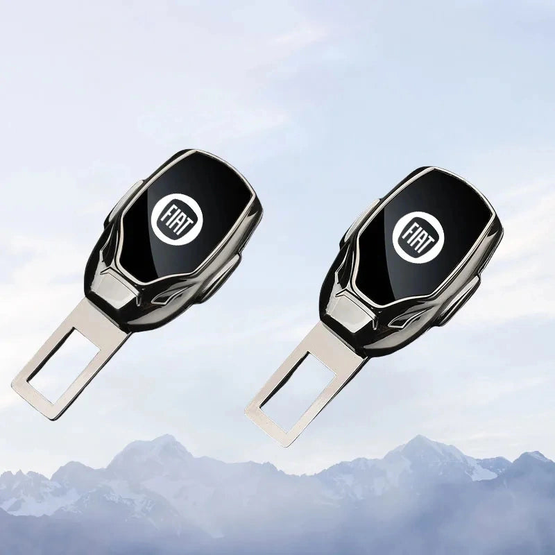 Two black and silver Fiat seatbelt key fobs with metal buckle inserts, shown against a soft blue sky and mountain backdrop.