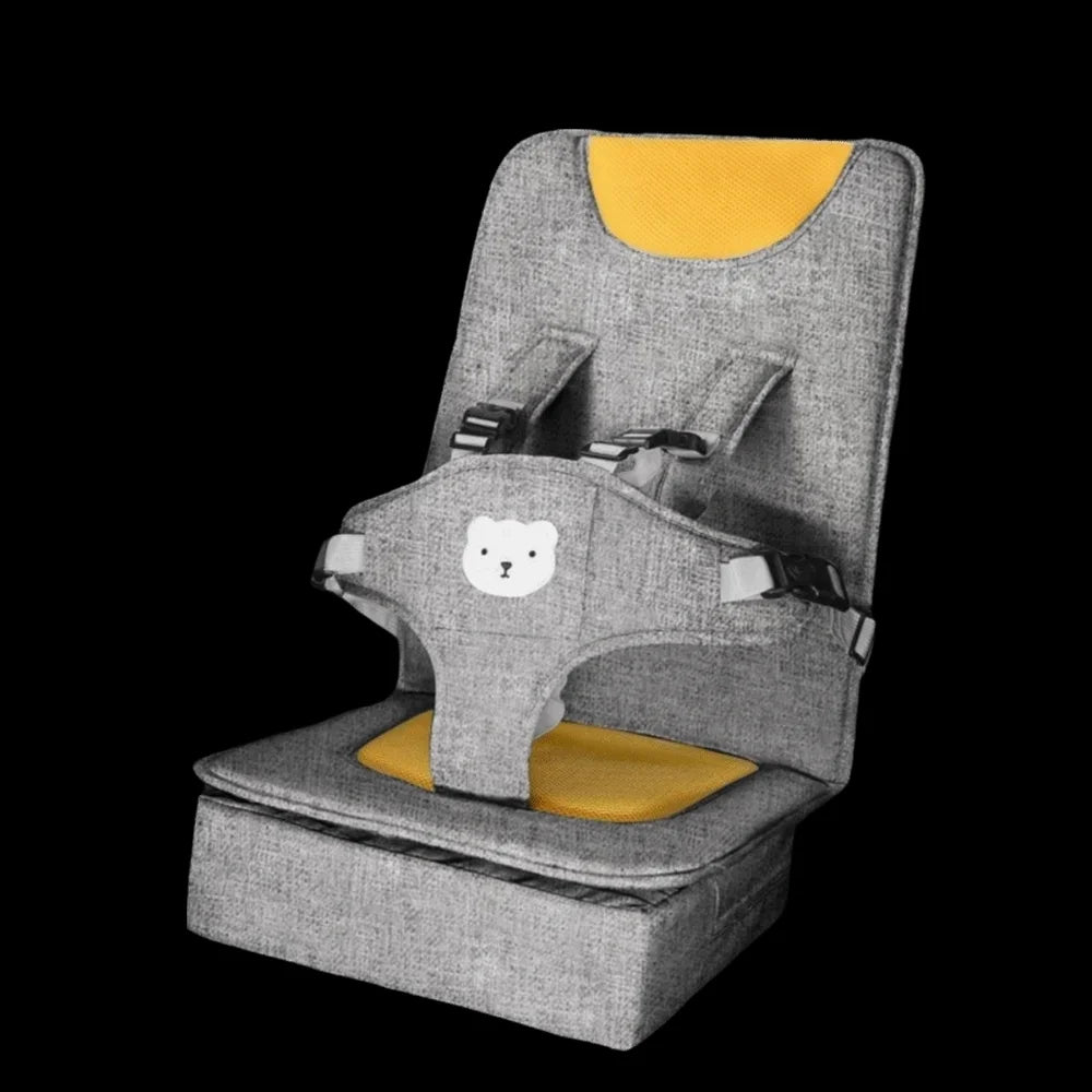 Front view of a beige portable baby booster seat with yellow accent padding and a teddy bear safety harness against a black background