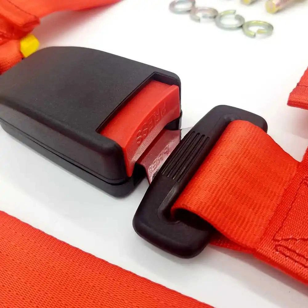 Close-up of a red 4-point racing harness showing the black press-button release buckle, steel latch tongue, and high-strength red nylon strap.